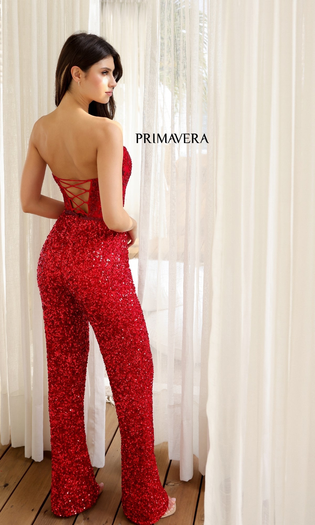 A woman stands near sheer curtains wearing the Long Prom Jumpsuit: Primavera 4383, featuring a red sequin design with a sheer corset bodice and lace-up back; “PRIMAVERA” is displayed in the background.